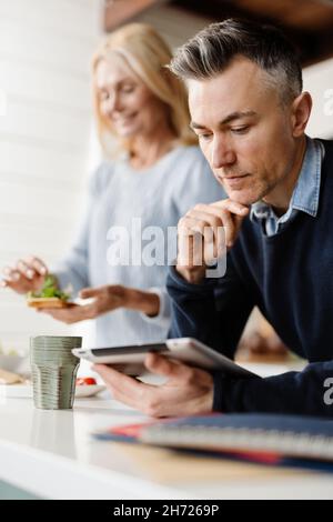 Ehepaar, Frau und Mann, die am Morgen vor der Arbeit frühstücken und an einem Tablet arbeiten Stockfoto