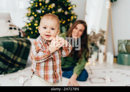 Ein kleines Kind hält einen Weihnachtsball. Im Hintergrund eine glückliche Mutter neben einem Weihnachtsbaum. Stockfoto