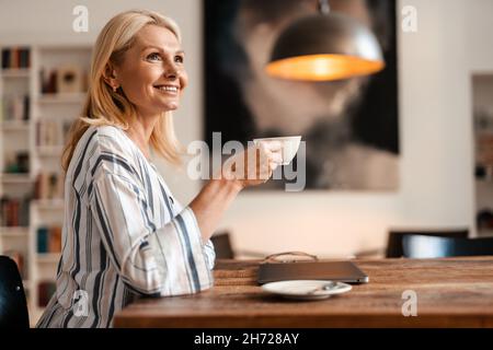 Blonde reife Frau lächelt und trinkt Tee, während sie zu Hause am Tisch sitzt Stockfoto