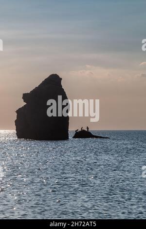 Drei Menschen werden gegen den frühen Abendhimmel auf einem Felsen neben dem Thurlestone, South Milton Sands, Devon, Großbritannien, silhouettiert. Stockfoto