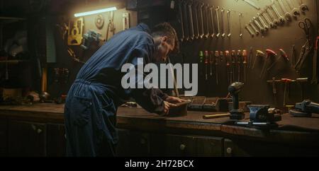 Schmied junger Mann in ehrfürchtigen Werkstatt Hintergrund er ein Stück Eisen oder Stahl in seiner Werkstatt, Weitwinkel-Rückansicht Schleifen Stockfoto