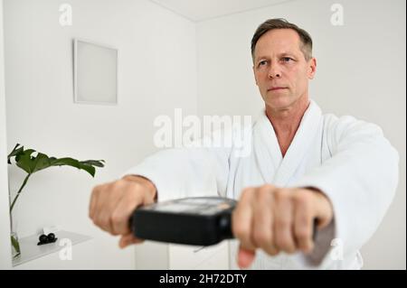 Porträt eines hübschen reifen 50 Jahre alten männlichen Patienten in weißem Frottee-Bademantel wiegen sich mit medizinischen Geräten - intelligente Waage, um bi zu bestimmen Stockfoto