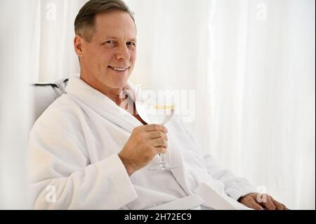 Schöner erfolgreicher reifer 50 Jahre alter europäischer Mann in weißem Frottee-Morgenmantel mit einem Glas Zitronenwasser und einem lächelnden, toothy Lächeln, das auf die Kamera schaut Stockfoto