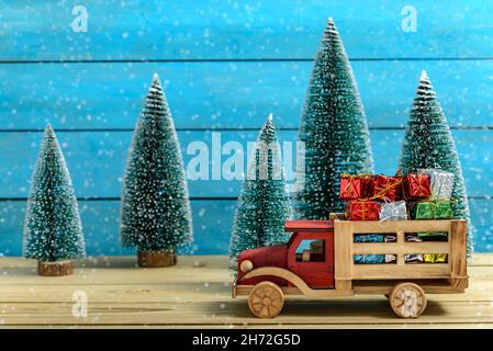 Weihnachtsgeschenke auf einem To Truck mit Weihnachtsmann-Hut neben Tannenbäumen, während es auf einem Holztisch schneit. Stockfoto