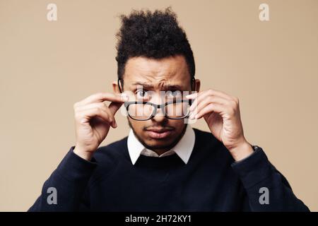 Junger besorgter Afro-Mann, der auf beigem Hintergrund über eine Brille schaut. Gesichtsausdruck, Emotionen Konzept Stockfoto