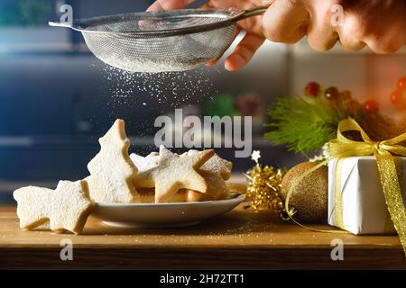 Mit einem Küchensieb hausgemachte Weihnachtskekse auf einem Teller auf der Küchenbank mit Puderzucker bestreuen. Vorderansicht. Horizontale Zusammensetzung. Stockfoto