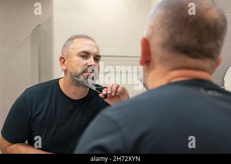 Porträt eines hübschen grauhaarigen Mannes in den 40er Jahren, der seinen Bart im Badezimmer selbst trimmt Stockfoto