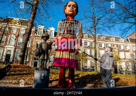 Den Haag, Niederlande. 19th. November 2021. Die riesige Marionette, die in der Nähe einer Skulptur eines kleinen Kindes posiert.am letzten Tag ihres Besuchs in den Niederlanden spazierte die riesige Marionette 'Little Amal', ein neun Jahre altes syrisches Flüchtlingsmädchen und über 11 Meter groß, durch das Stadtzentrum von Den Haag. Wo sich das Repräsentantenhaus befindet, um mit den Menschen auf den Straßen zu interagieren. Dieser Besuch wurde von Amare organisiert, einem neuen kulturellen Zentrum im Rahmen des Open Festivals. Mit ihrem Besuch will Amal in ganz Europa die Aufmerksamkeit auf die Not junger Flüchtlinge lenken, die aus Syrien geflohen sind. Während ihres VI Stockfoto