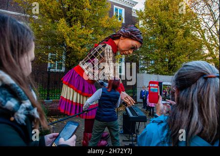 Den Haag, Niederlande. 19th. November 2021. Die kleine Amal sah, wie sie versuchte, ein Klavier zu spielen.am letzten Tag ihres Besuchs in den Niederlanden spazierte die riesige Marionette "Little Amal", ein neunjähriges syrisches Flüchtlingsmädchen und über 11 Meter groß, durch das Stadtzentrum von Den Haag, wo sich das Repräsentantenhaus befindet, Mit den Menschen auf der Straße zu interagieren. Dieser Besuch wurde von Amare organisiert, einem neuen kulturellen Zentrum im Rahmen des Open Festivals. Mit ihrem Besuch will Amal in ganz Europa die Aufmerksamkeit auf die Not junger Flüchtlinge lenken, die aus Syrien geflohen sind. Während ihres Besuchs im Stadtzentrum, am Stockfoto