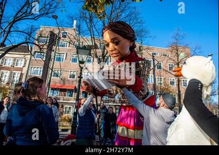 Den Haag, Niederlande. 19th. November 2021. Zwei Mädchen werden gesehen, wie sie der kleinen Amal ein Studentencarnet von der Universität Leiden geben.am letzten Tag ihres Besuchs in den Niederlanden spazierte die riesige Marionette 'Little Amal', ein neun Jahre altes syrisches Flüchtlingsmädchen und über 11 Meter groß, durch das Stadtzentrum von Den Haag, Wo sich das Repräsentantenhaus befindet, um mit den Menschen auf den Straßen zu interagieren. Dieser Besuch wurde von Amare organisiert, einem neuen kulturellen Zentrum im Rahmen des Open Festivals. Mit ihrem Besuch will Amal in ganz Europa die Aufmerksamkeit auf die Not junger Flüchtlinge lenken, die aus Süd geflohen sind Stockfoto