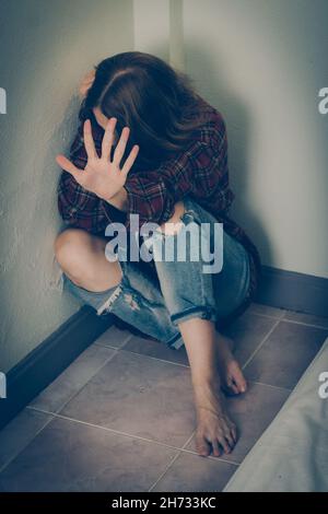 Eine Frau in Angst sitzt auf dem Boden gegen die Wand und bedeckt ihr Gesicht Stockfoto