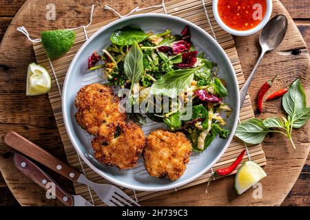 Köstliche Thai-Fischkuchen mit asiatischem Salat und süßer Chilisauce auf einer rustikalen Holzplatte. Stockfoto