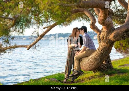 Junges biraziales Paar, das auf einem großen Baumzweig neben dem Wasser sitzt und sich zum Kuss anlehnt Stockfoto