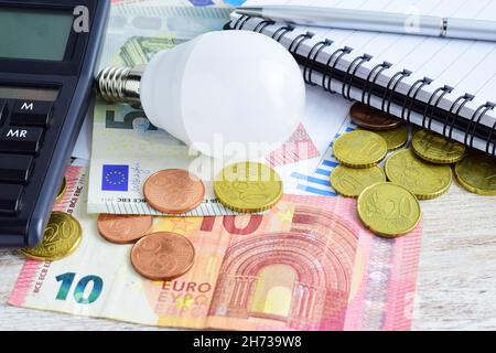 Glühbirne, Euromünzen, Banknoten, Taschenrechner und Notizbuch auf dem Holztisch Stockfoto