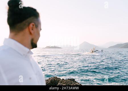 Groom steht auf den Felsen und schaut auf die Motoryacht, die auf dem Meer segelt Stockfoto