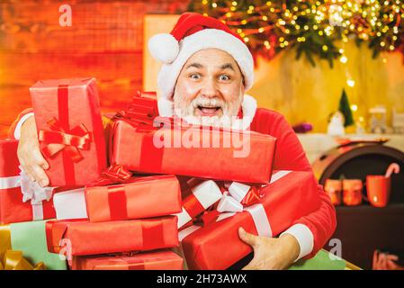 Aufgeregter Weihnachtsmann mit Weihnachtsgeschenk. Weihnachtskarte mit Weihnachtsmann. Großvater mit einem weißen Bart, der auf dem weihnachtlichen Hintergrund posiert. Stockfoto