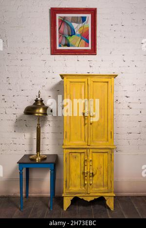 Still Life von Vintage gelben Schrank, gerahmte Gemälde, Messing Tischlampe und Vintage kleinen Tisch auf dunklem Holzparkett Boden und weißen Ziegelwänden in Studio Stockfoto