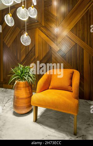 Moderner orangefarbener Sessel, hoher Holztoper mit grünen Büschen und moderner Kronleuchter aus Glas in einem Flur mit dekorierter Holzverkleidung und weißem Marmorboden Stockfoto