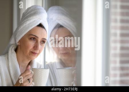 Moody Foto einer Frau mittleren Alters mit einem Handtuch-Turban auf dem Kopf während ihrer morgendlichen Routine, mit einer Tasse Kaffee in den Händen, die sich auf den Windo lehnt Stockfoto