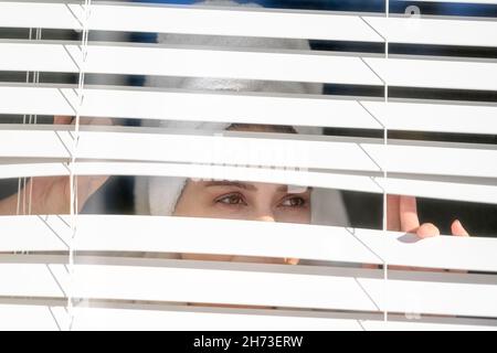 Eine Frau mittleren Alters mit einem Handtuch-Turban auf dem Kopf zieht den horizontalen Blind weg und schaut durch das Fenster, um zu sehen, was vor sich geht oder was zu sehen ist Stockfoto