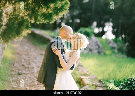 Bräutigam in einem schwarzen Anzug umarmt und küsst die Braut in einem weißen Kleid und steht auf einem Pfad im Park vor dem Hintergrund von grünen Bäumen Stockfoto