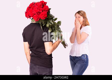 Kuriermann in schwarzer Uniform lieferte einer verblüfften blonden Frau einen wundervollen Strauß roter Rosen, sie kann es nicht glauben und bedeckt ihr Gesicht mit ihr Stockfoto