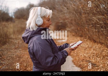 Im Herbstpark der Stadt hört eine schöne blonde Frau im mittleren Alter mit Kopfhörern Musik. Stockfoto