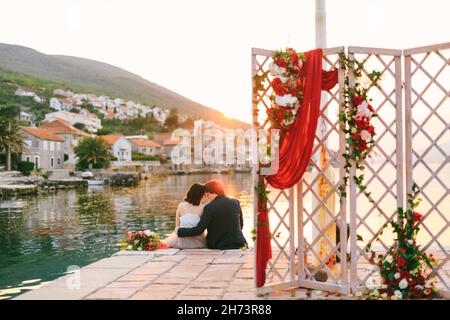 Das Brautpaar sitzt umarmt sich am Pier vor dem Hintergrund des Wassers, der Berge und der alten Häuser. Im Vordergrund befindet sich ein durchbrochener Bildschirm Stockfoto