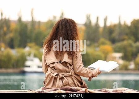 Junge afroamerikanische Frau mit geöffnetem Buch in der Nähe des Flusses Stockfoto