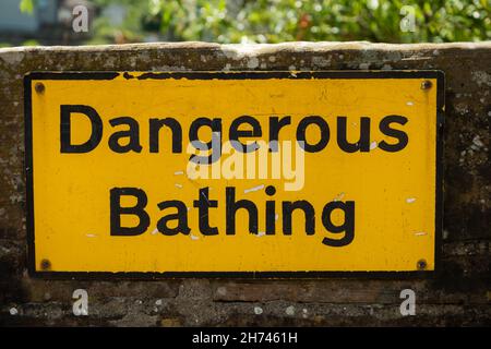 Ein gelbes Warnschild für gefährliches Baden, das an einer Steinmauer neben dem Wasser befestigt ist Stockfoto