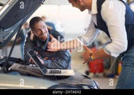 Lächelnder Mechaniker mit Laptop im Auto. Professioneller Arbeiter repariert Auto Stockfoto