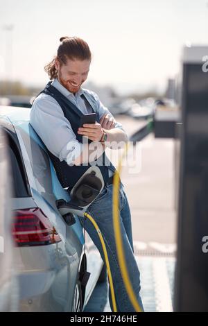 Junger Mann mit Smartphone in der Nähe eines Elektroautos an der Ladestation Stockfoto