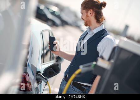 Ein junger lächelnder Mann hält ein Telefon, während das Elektroauto an der Ladestation aufgeladen wird Stockfoto
