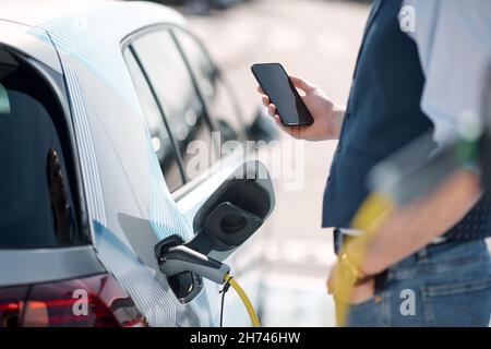 Der junge Mann hält ein Telefon, während das Elektroauto an der Ladestation aufgeladen wird Stockfoto