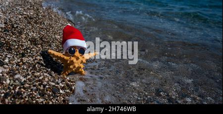 Seesterne in weihnachtsmannhut und Sonnenbrille an einem Kiesstrand am Meer. Stockfoto