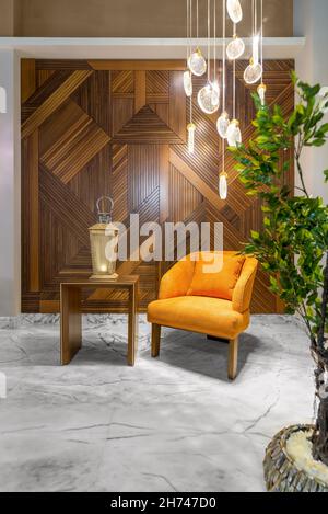 Moderner orangefarbener Sessel, beleuchtete Holzlaterne auf einem kleinen Holztisch und moderner Kronleuchter aus Glas in einem Flur mit dekorierter Holzverkleidung und weißem Marmorfußboden Stockfoto