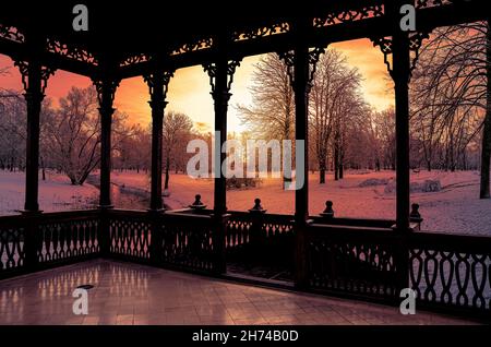 Blick auf den Sonnenuntergang über dem gefrorenen Fluss und dem verschneiten Wald vom Holzbalkon. Hölzerne durchbrochene Galerie oder Pavillon. Stockfoto