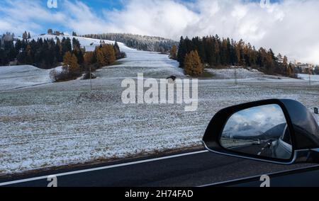 Eine Aufnahme aus dem Fahrerfenster eines Elektroautos mit schneebedeckten alpen-Bergen. Kalter, wolkiger Herbsttag. POV erste Person Ansicht auf einem geschossen Stockfoto