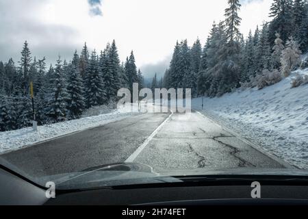 Ein Schuss auf die Bewegung hinter der Windschutzscheibe eines Elektroautos mit schneebedeckten alpen-Bergen. Kalter, wolkiger Herbsttag. POV-Ansicht der ersten Person aufgenommen Stockfoto