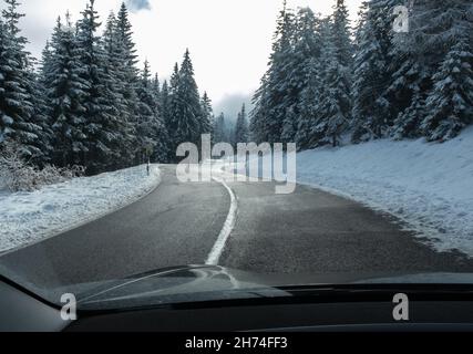 Ein Schuss auf die Bewegung hinter der Windschutzscheibe eines Elektroautos mit schneebedeckten alpen-Bergen. Kalter, wolkiger Herbsttag. POV-Ansicht der ersten Person aufgenommen Stockfoto