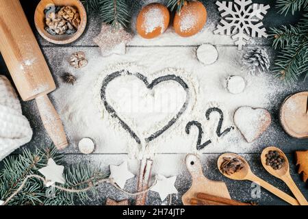 Herz und Zahlen 22 geschrieben auf Mehl auf schwarzem Tisch mit Zweigen von Weihnachtsbaum, Backzubehör und Zutaten bestreut. Frohe Weihnachten und Stockfoto