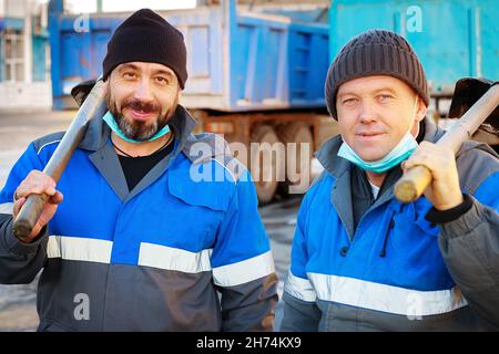 Zwei Arbeiter in Arbeitskleidung und medizinischen Masken stehen mit Schaufeln auf der Straße und blicken in die Kamera. Portrait der kaukasischen Handwerker. Männliche Handwerker auf der Suche nach einem Nebenjob Stockfoto