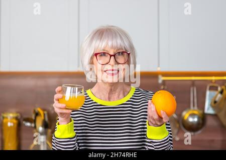 Fröhliche, ziemlich ältere Frau in gestreiftem Pullover, die Orange hält und Orangensaft trinkt, während sie in der Küche steht. Gesunde, saftige Lebensmacht Stockfoto