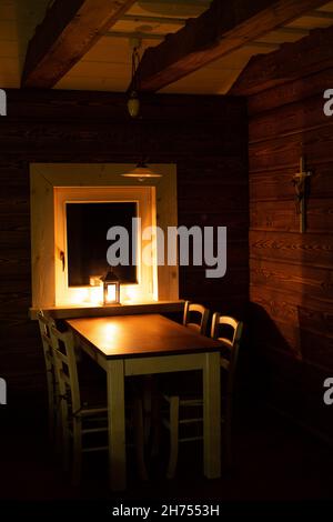 Holztisch, Stühle, Laterne im Fenster, warmes Kerzenlicht, Abend in einem einfachen Holzhaus, Holzfenster, Holzhaus im alten Stil Stockfoto