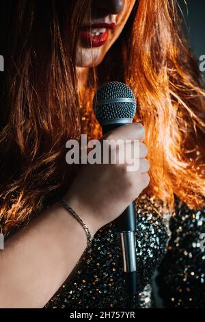 Gesichtsloses Porträt einer Rotschopf-Frau in glitzerndem Abendkleid mit Mikrofon auf dunklem nächtlichen Hintergrund. Nicht erkennbare Sängerin mit Modern Stockfoto