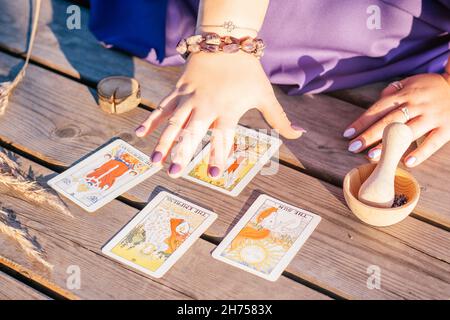 Die Frau hält ihre Hand mit violetten Nägeln über vier Tarot-Karten, die auf einer Holzfläche neben Stacheletts und Lavendel verteilt sind. Minsk, Weißrussland - 07.28.2021 Stockfoto