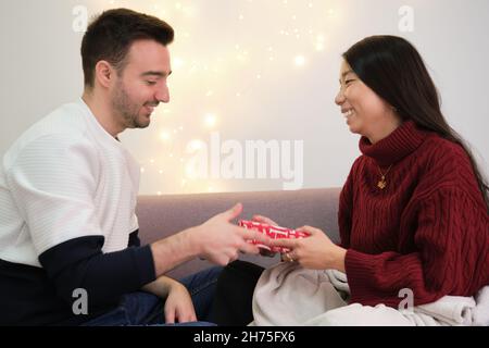 Junge asiatische Frau, die seinem kaukasischen Freund ein Weihnachtsgeschenk macht. Stockfoto