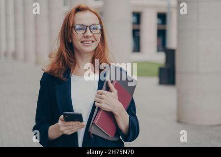 Fröhliche rothaarige Geschäftsfrau in offizieller Kleidung, die im Freien steht und das Mobiltelefon benutzt Stockfoto