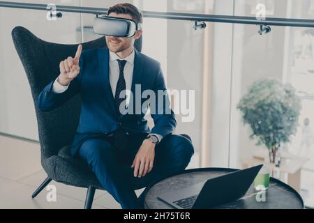 Lächelnder Geschäftsmann mit VR-Headset-Brille, während er in der Bürolobby sitzt und am Laptop arbeitet Stockfoto