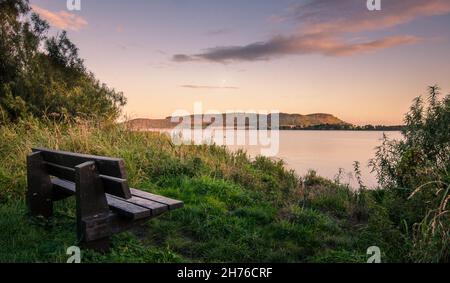 Eine Holzbank mit Blick auf den Sonnenuntergang auf das ruhige Wasser des Sees Stockfoto